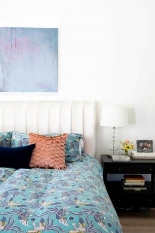Ein weißes Schlafzimmer mit farbenfroher blauer Bettwäsche mit Vogelmuster, einem Stoffkissen mit Schuppenmuster und einem Nachttisch