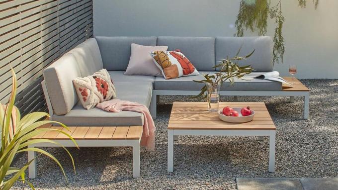 Un sofá de esquina al aire libre con cojines grises y una mesa de centro con paneles de madera y estantes laterales