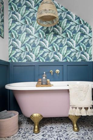 Salle de bains lambrissée bleue avec papier peint à motifs palmiers, carrelage à motifs et baignoire autoportante rose