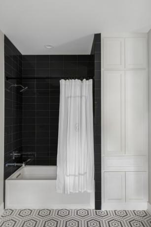 검정, 흰색 및 회색 욕실에는 검은색 타일의 샤워 시설, 샤워기가 설치된 욕조, 흰색 샤워 커튼, 무늬가 있는 바닥 타일이 있습니다.