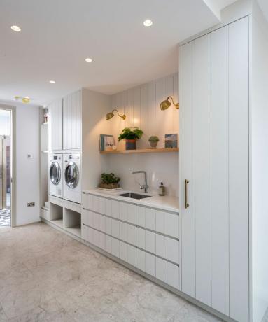 Dolaplara yerleşik çamaşır kurutma makinesi, saksı bitkileri, yardımcı lavabo ve pirinç duvar lambaları ile aydınlık ve beyaz çamaşır odası