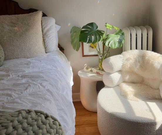 Hygge-Schlafzimmer mit einer weißen Bettdecke, einem grünen Überwurf, Pflanzen und einem flauschigen weißen Stuhl