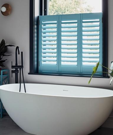 Baignoire blanche dans la petite salle de bains avec volets bleus et cadre de fenêtre noir