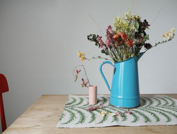 bir masada kuru çiçeklerle dolu mavi sürahi, yanında küçük pembe vazo, solda kırmızı sandalye