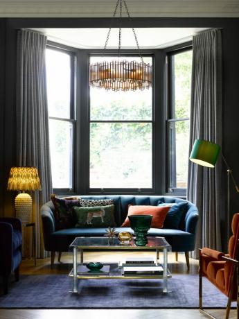 ruang tamu dicat abu-abu dengan jendela ceruk, sofa dan kursi biru, kursi oranye, lampu lantai, lampu meja, permadani biru, lampu gantung modern, meja kopi kaca