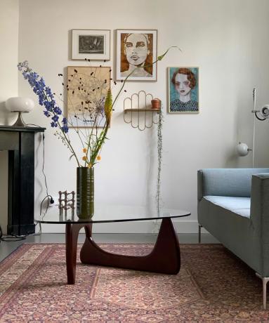 MCM-lounge met sculpturaal glas en donkerhouten salontafel, een vaas met hoge bloemstengels en galerijwandmontage.