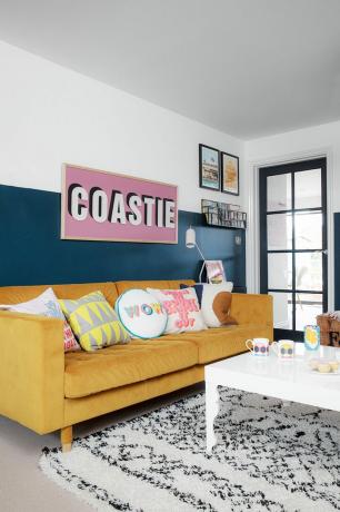 Kék nappali monokróm Scandi szőnyeggel, fehér dohányzóasztallal, sárga kanapéval és nagy 