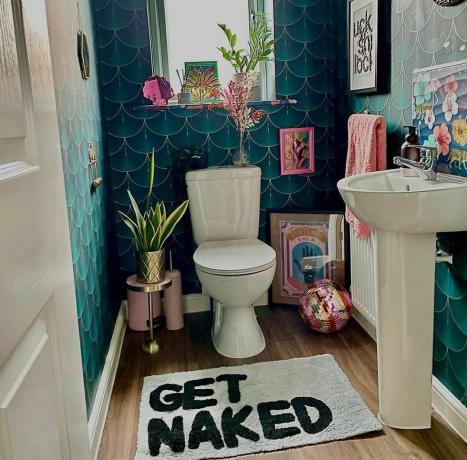 Ein Badezimmer mit blaugrün gefliesten Wänden und einer Badematte mit der Aufschrift „Zieh dich aus“
