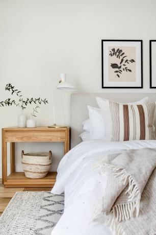 neutralus miegamasis su medine lova, tekstūruota balta ir avižinių dribsnių patalyne ir kilimėliu, krepšeliu, balta staline lempa, meno kūriniais
