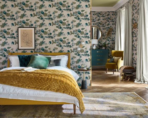 Een slaapkamer met geometrisch en botanisch patroonbehang, groot mosterdkleurig bedframe, bed met mosterdgele sprei, ingelijste muurkunst boven bed en groot vloerkleed op houten vloer