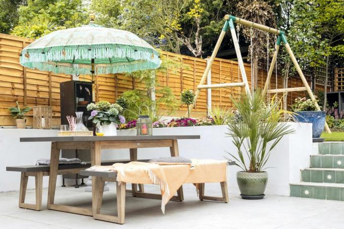 فناء ببلاط رمادي فاتح ومجموعة مقاعد خشبية لتناول الطعام ومظلة خضراء ودرجات من البلاط إلى الحديقة