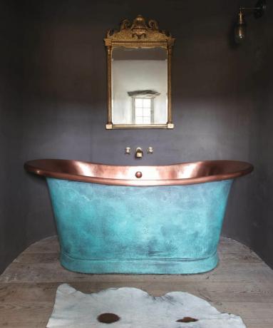 Ванная комната с темной краской на стенах, искусственным ковриком с животным принтом и медной ванной, окрашенной синей краской, с витиеватым латунным зеркальным дизайном.