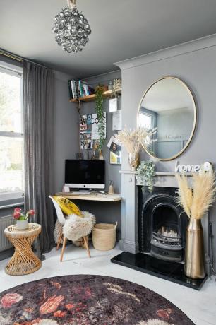 Namų biuras su pilkomis sienomis, baltai dažytomis medinėmis grindimis, purpurinio rašto kilimėliu, tradiciniu židiniu ir apvaliu veidrodžiu