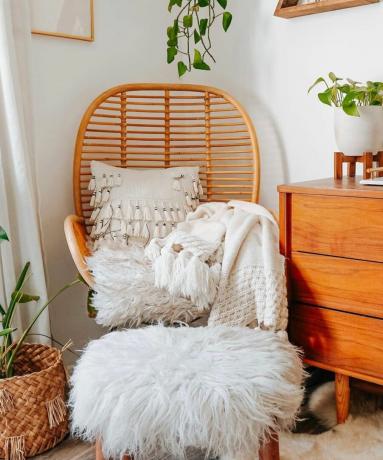 Een geweven stoel met een wit kussen en plaids, plus planten eromheen