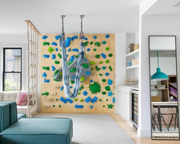 Uma sala de estar com parede de escalada feita de madeira e pegas de cor verde-azul, rede suspensa no teto e dispositivo de escalada por corda