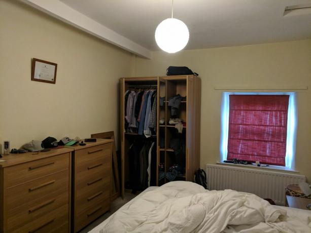 Fotografie „Înainte” a dormitorului cu pereți crem, comodă din lemn și jaluzele din material roșu