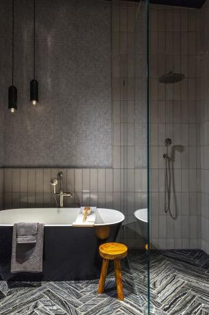 banheiro com áreas de banheira e chuveiro divididasparede de vidro, paredes cinza, azulejos cinza claro, azulejos espinha de peixe, azul preto, banquinho, toalhas, pingente