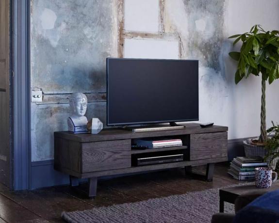John Lewis & Partners Calia TV Stand i mørk stue med brunt tæppe, bare vægge og plante