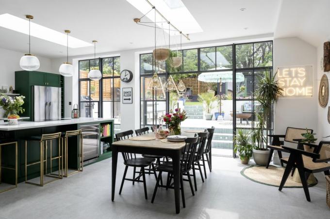 Pramoninio stiliaus virtuvė-valgomasis su betoninėmis grindimis, žalia virtuve ir sala, juodu mediniu stalu ir kėdėmis bei svetainės zona su rotango kėdėmis ir neonine šviesa „likime namuose“