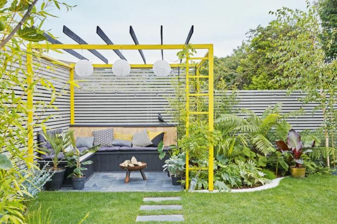 Trooppinen puutarhaistutus istuinalueella ja keltaiseksi maalattu pergola
