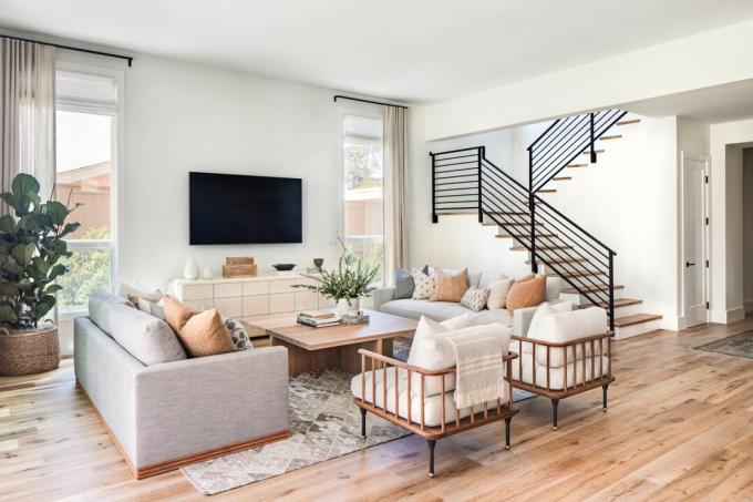 harmaa ja beige neutraali olohuone, jossa yhteensopivat sohvat, puinen neliönmuotoinen sohvapöytä, kaksi puista nojatuolia, portaikko, parkettilattia, kasveja, harmaa matto