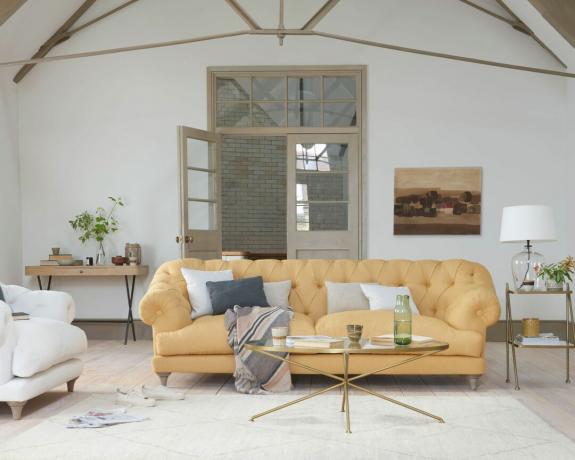 Modernes Wohnzimmer im Landhausstil mit weichem gelbem Sofa mit Knopfleiste, weißen Wänden und warmgrau gestrichenen Türen und Dachkonstruktionen.