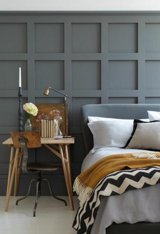 Ein getäfeltes, grau gestrichenes Schlafzimmer mit grauem Kopfteil auf dem Bett mit senffarbenem und gestreiftem Überwurfdekor