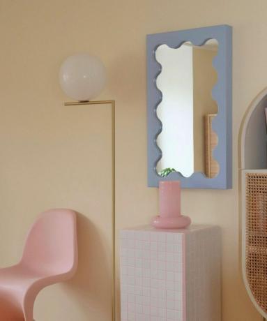 zils viļņojošs spogulis istabā ar rozā krēslu, zelta lampu un baltu flīžu sānu galdu