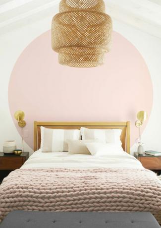 Cerchio rosa dipinto con cornici letto in legno con biancheria da letto spogliata e tiro a maglia grosso rosa. Il lampadario intrecciato centra lo spazio.