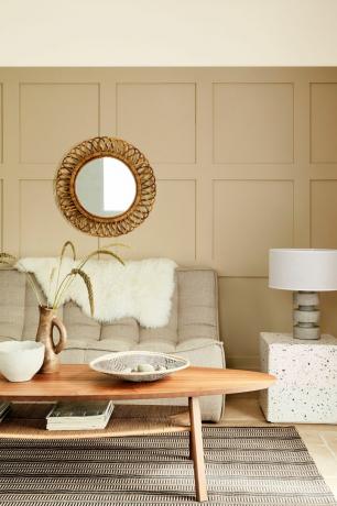 neutraali olohuone beige-, kivi- ja okraväreillä, puinen ovaali sohvapöytä, pyöreä rottinkipeili, panelointi, moderni sohva, kivilattia, kuvioitu matto, käsityöläistarvikkeet