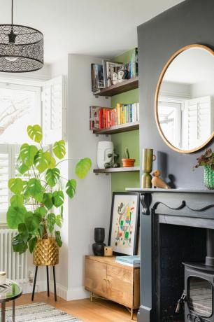 Bücherregal in grüner Nische im Wohnzimmer, mit übergroßem Monstera in Gold-Pflanzgefäß mit Beinen