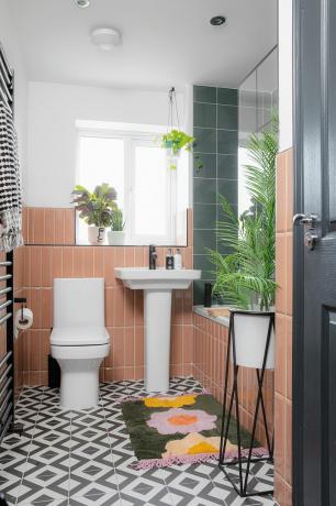 분홍색 세로형 지하철 벽 타일, 단색 패턴 바닥 타일, 흰색 스위트룸 및 녹색, 분홍색 및 노란색 꽃무늬 욕실 매트가 있는 작은 욕실