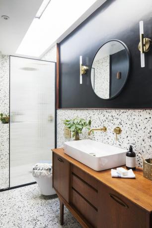 Badeværelse med terrazzo væg- og gulvfliser, et segment af vægmalet sort, ribbet glas bruseafskærmning, teak skænk drejet vaskeskab, messing hardware og rundt spejl