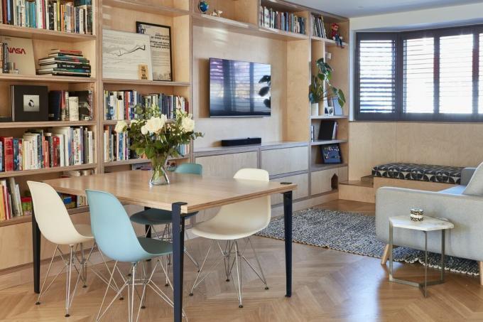 Vanerista valmistettu olohuone, jossa on sisäänrakennettu säilytystila, joka johtaa avoimeen ruokailutilaan, jossa on Eames-tyyliset tuolit