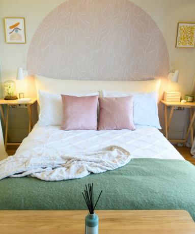 白いシーツ、ピンクのクッション、テクスチャーのあるグリーンのクッションを備えたベッド。ヘッドボードの上には落ち着いたグラフィックのピンク色で描かれた半円形があり、両側に白い壁取り付け用燭台が付いています。