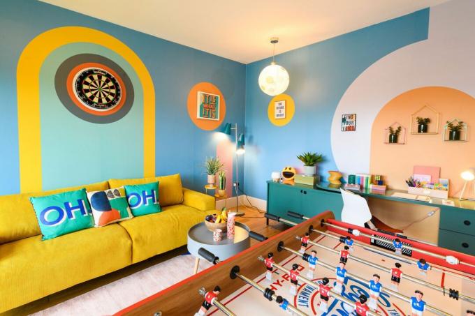 Ljust färgat rum med blå, gula och orange accenter, en bordsfotboll, gul soffa och grafiska kuddar