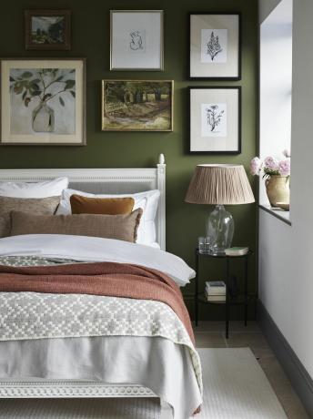 groene slaapkamer met galerijwand, rustiek gevoel, wit geverfd bed, okergele deken en kussen, lamp op glasbasis, linnen kussens, crèmekleurig vloerkleed