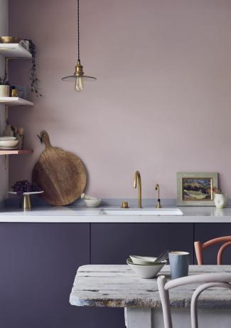 Küche mit dunkelvioletten Schränken, rosafarbener Wand, weißer Arbeitsplatte, offenen Regalen, Messingarmaturen, rustikalem Tisch