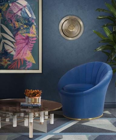 Salon bleu MCM audacieux avec chaise courbe en velours bleu, table basse ronde en métal, murs bleu foncé et imprimé de feuilles multicolores sur le mur.
