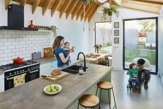 Et kjøkken med stor svingdør og bildevindu, benkeplater og gulv i betong-effekt og synlige bjelker i det forlengede taket