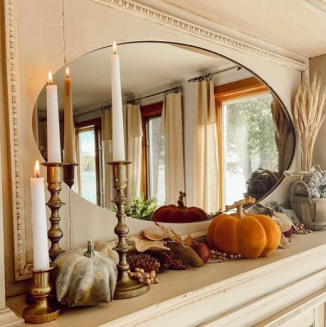 Un manteau de cheminée avec miroir ovale, citrouilles, herbe de la pampa dans un vase, chandeliers, pommes de pin et feuillage d'automne