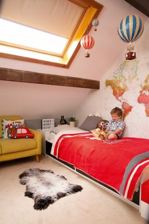 Kamar tidur anak dengan mural dinding peta, bed cover merah putih dan kursi berlengan kuning