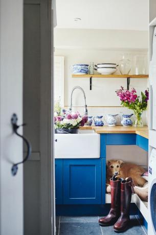青いキッチンユニット、オープンシェルフ、ベルファストシンク、木製の調理台を備えたユーティリティルームのベッドにいる犬