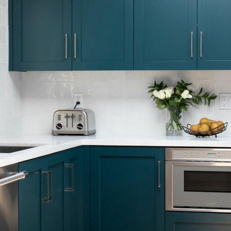 Virtuvės erdvė su mėlynos spalvos spintelėmis, baltais stalviršiais ir atlošomis