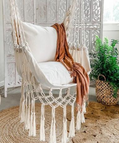 Un fauteuil suspendu en macramé avec un tapis en jute en dessous et une plante à côté