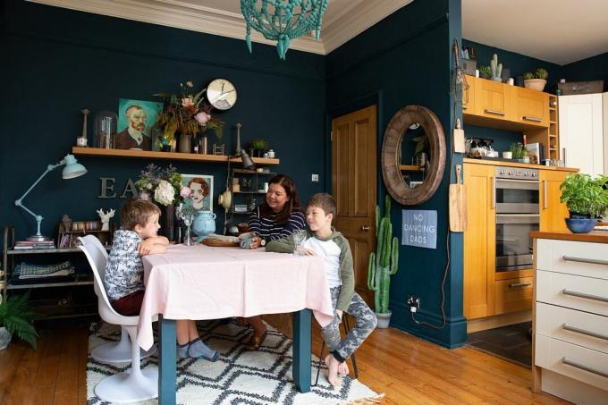 ครอบครัวนั่งรอบโต๊ะอาหาร ห้องมีผนังสีน้ำเงินเข้มและมีโคมระย้าสีเทอร์ควอยซ์ห้อยลงมาจากเพดาน โต๊ะสีเทาปูด้วยผ้าปูโต๊ะสีชมพูอ่อน