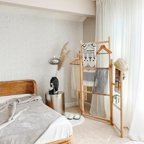 Pembatas ruangan bambu digantung dengan selimut dan pakaian bermotif mono di kamar tidur netral dengan wallpaper geo halus, dan tekstur alami.