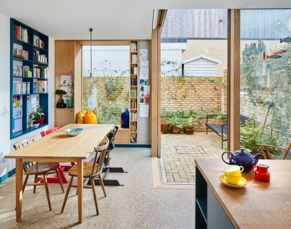 Ampliación de la cocina-comedor con mesa de comedor y sillas de madera, asiento junto a la ventana, estanterías empotradas pintadas de azul y puertas corredizas de esquina de madera que dan al jardín del patio