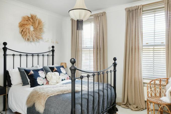 Camera da letto con pareti bianche, tende beige, giroletto in metallo nero, cuscini floreali blu e ghirlanda di fiori secchi