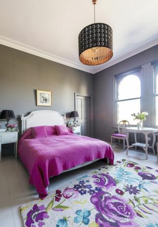 Soveværelse med slående lyserød seng, hvidt sengegavl, blomstret tæppe og dekorativ lampeskærm i kobbereffekt